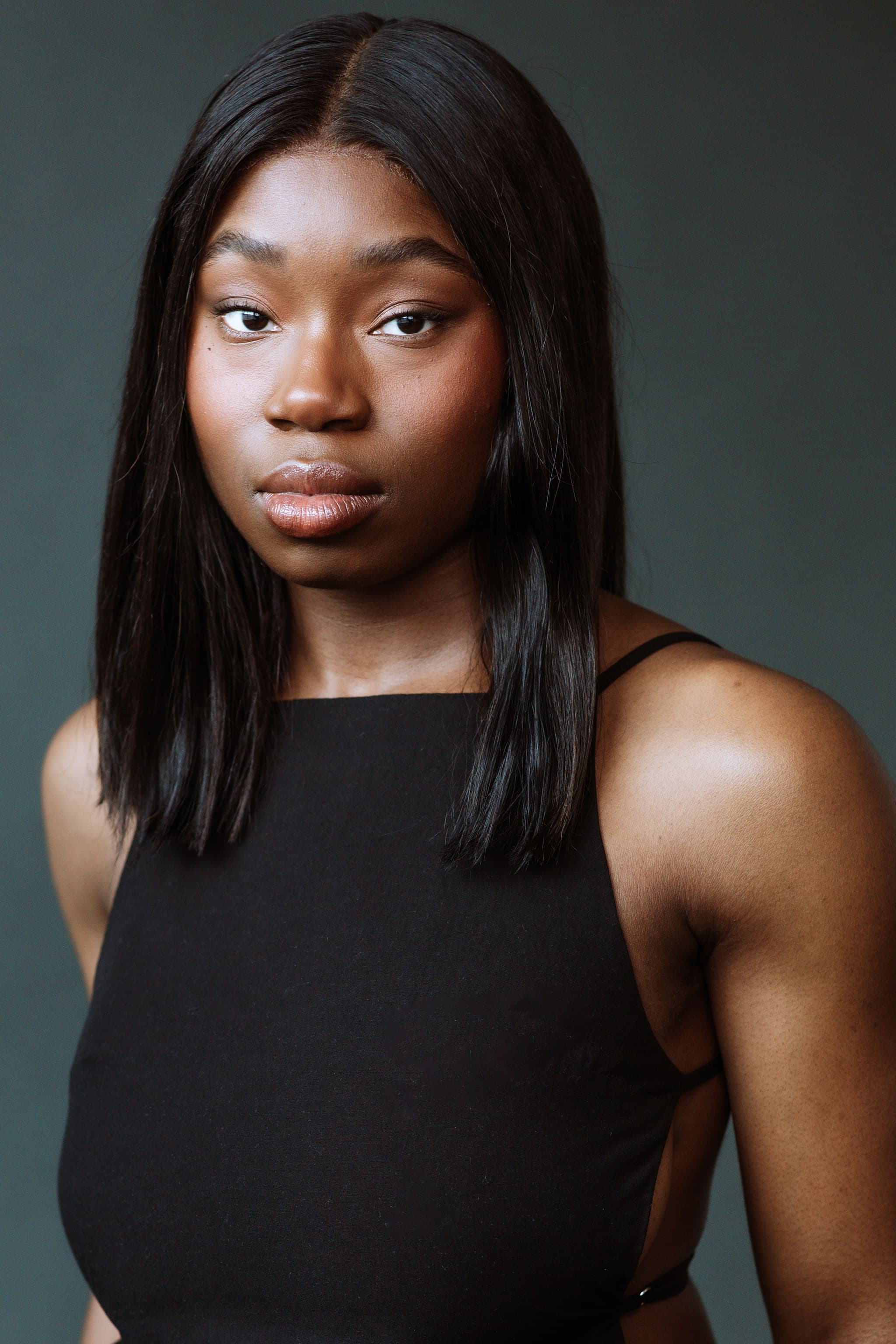 Cassandra a Black woman with shoulder length straight black hair soflty glancing, in front of charcoal grey backdrop, wearing camouflage pants and black top with two thin straps.