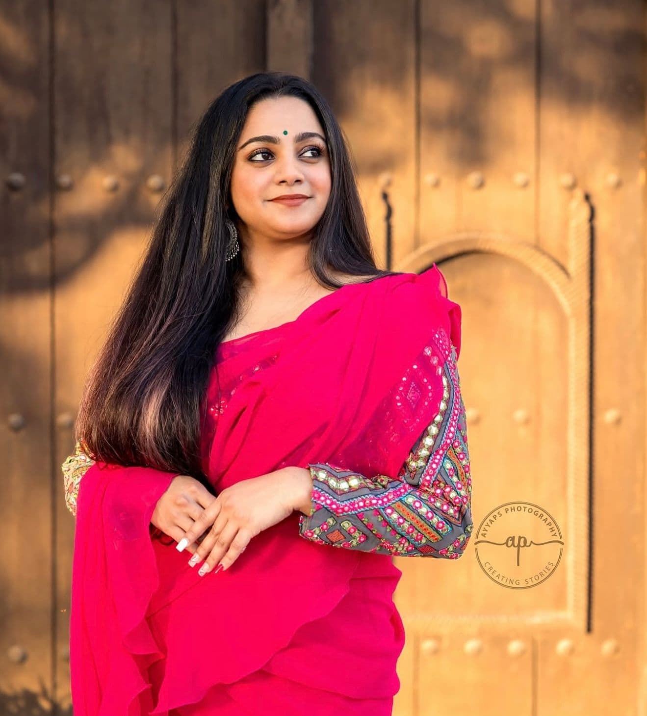 Arathi, an Indian girl is wearing a bright pink saree with an intricately embroidered blouse. Her long, dark hair is left open, and she has styled her look with a small green bindi, statement earrings, and a natural makeup with smile on her face.