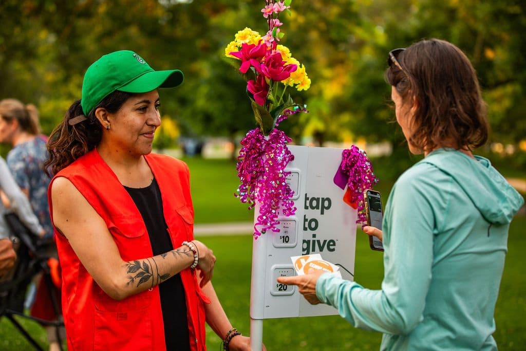 Photo of a volunteer in an orange vest and a green hat greeting a patron in an teal seater. She is holding a white machine decorated with flowers and strings.