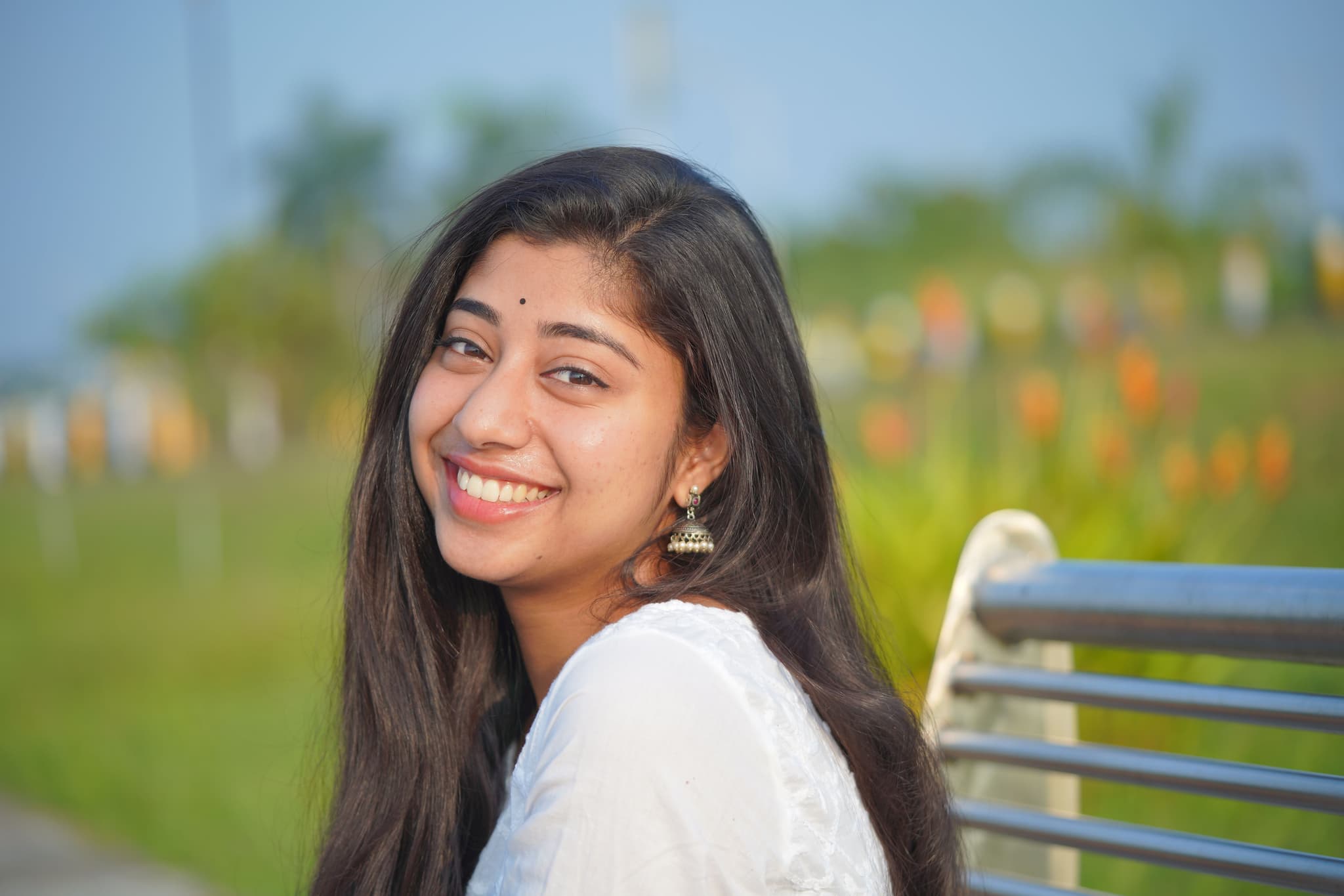 Roweena, a South Asian woman with long straight hair, smiles warmly in a color photo. She is wearing a white embroidered outfit and traditional earrings, turning slightly to face the camera.