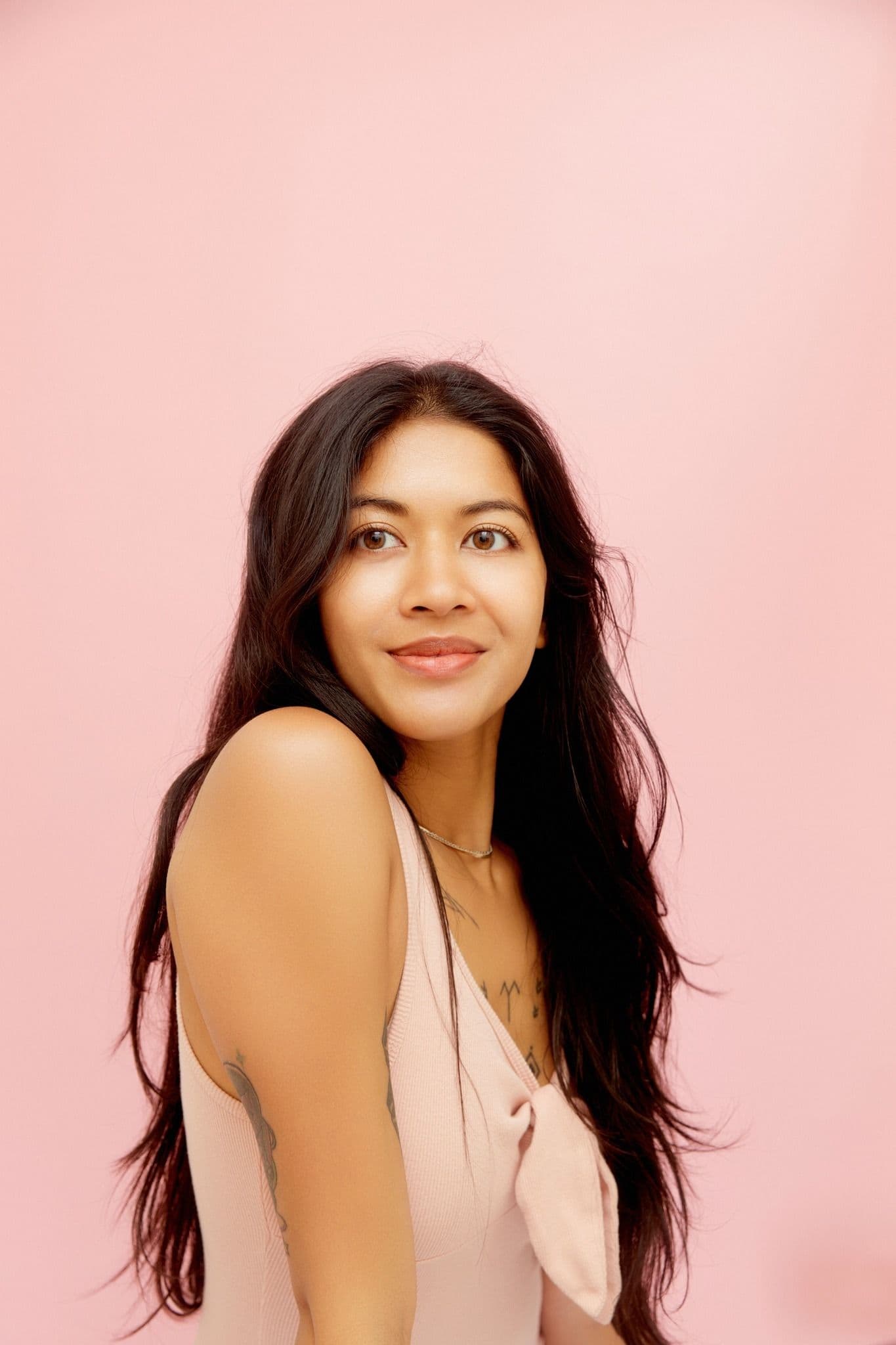 Candace Kumar, a Filipino Indo-Fijian woman with long wavy black hair, gently smiles and gazes toward the left against a pink backdrop. She is wearing a light pink tank top with a bow and a gold necklace.