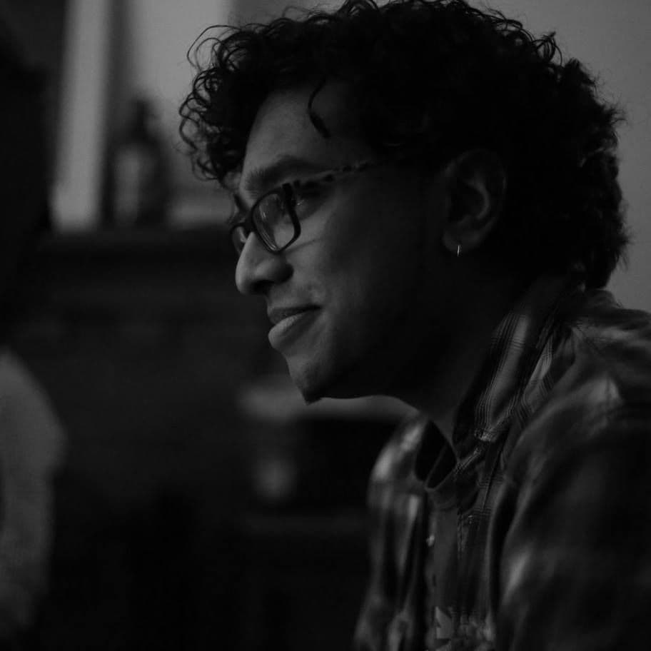 Chin, a South Asian man with short curly hair is seen in profile, smiling in a black and white photo. He has thick framed glasses and an earring. He is wearing a plaid long-sleeved shirt on top of a graphic tee.