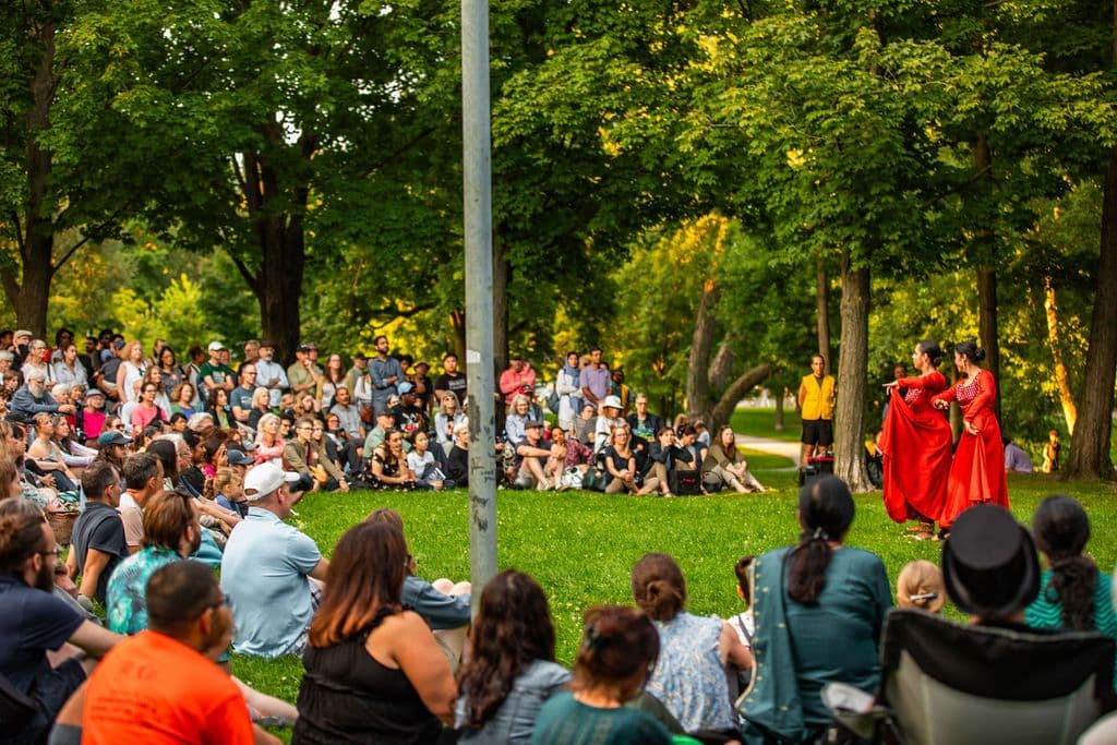 Two dancers in long flowy red dresses and hair tied in buns perform in the middle of a grassy field surrounded by trees. Many audience members are seated and standing in a semi circle watching them.