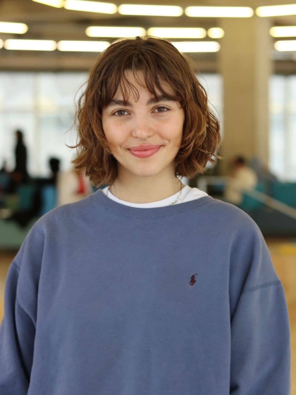 Bella, a white woman with shoulder length brown hair, smiles at the camera. She is wearing a light purple sweater.
