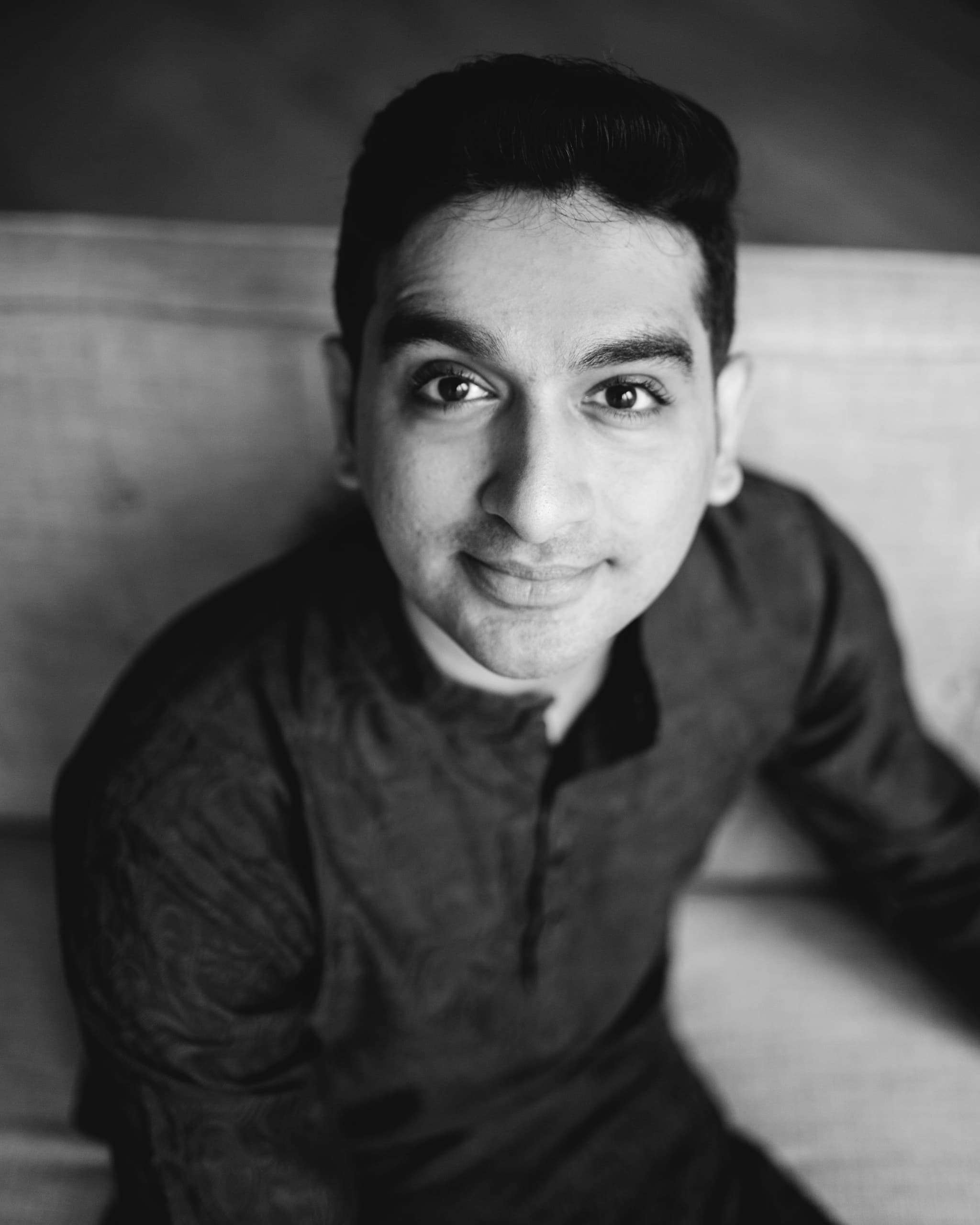 Hari, an Indian man, smiles softly to the camera while glancing upwards. He is wearing a high-collar black button down shirt.