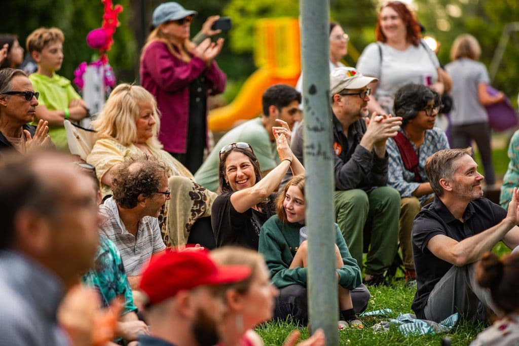 A crowd seated and standing in the grass. Many are smiling and have their hands raised clapping.