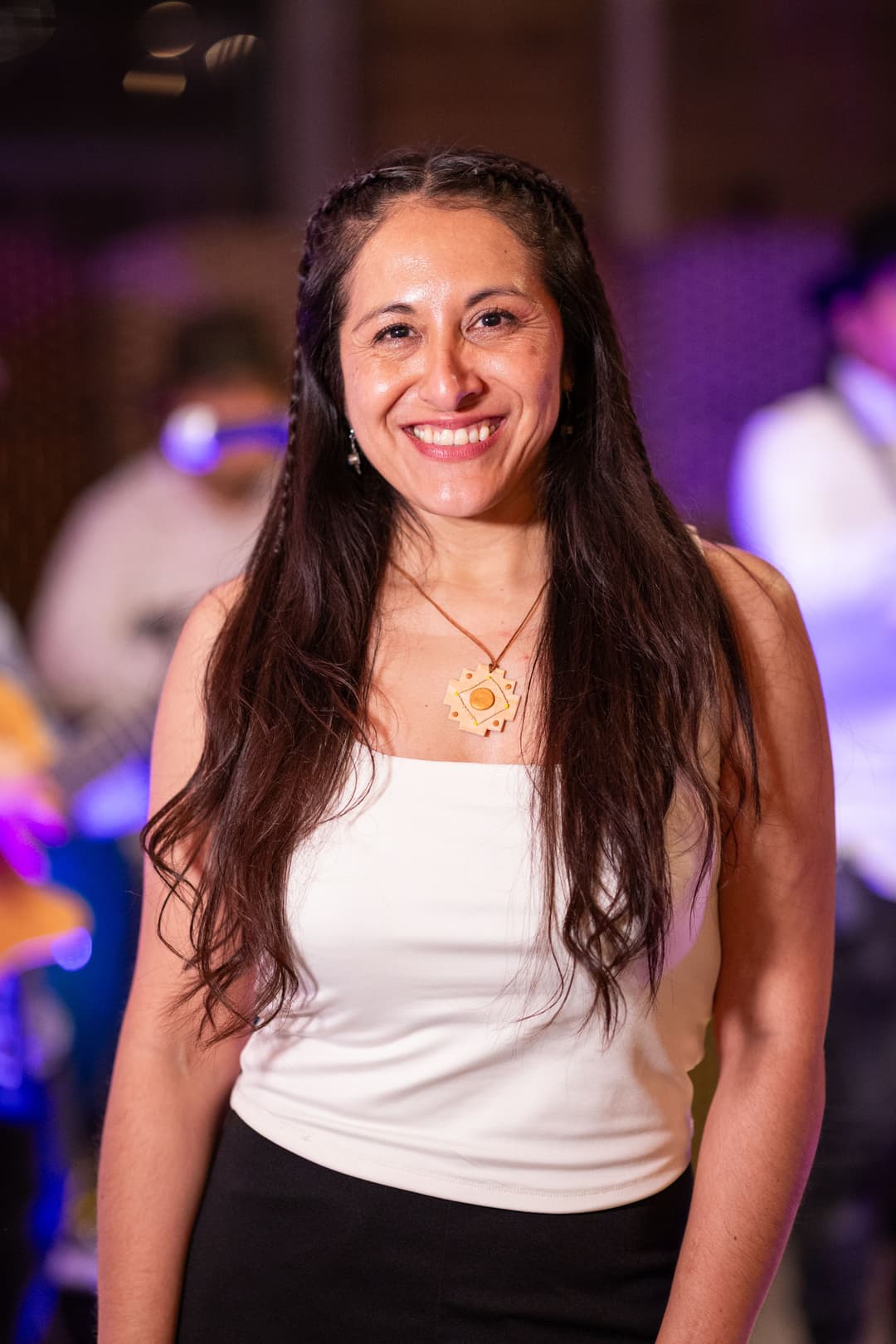 photo of a latina woman with long, dark hair with two small braids. She wears a white top and a yellow pendant necklace.