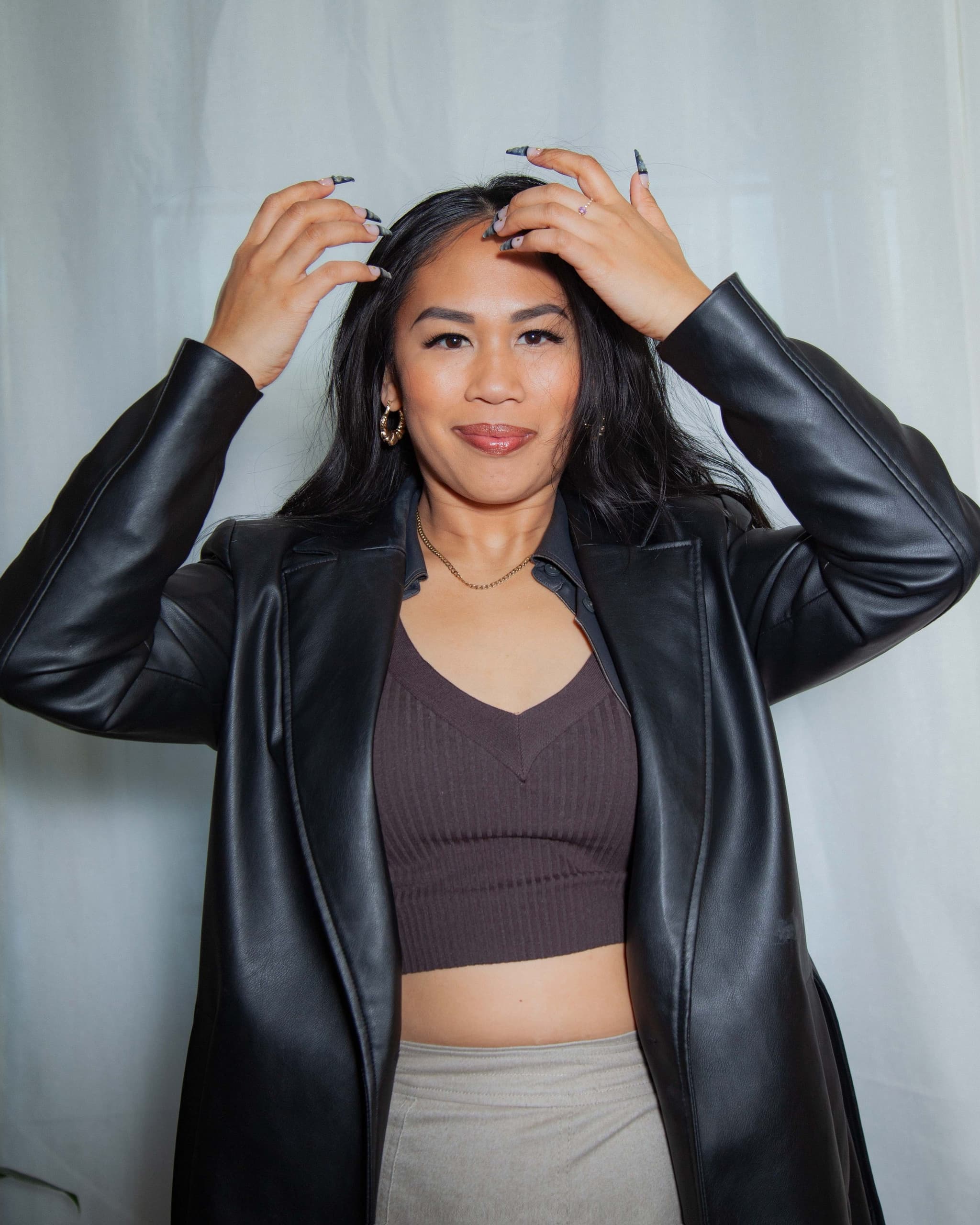 Danah, a filipinx with black hair, smiling while parting her hair in a black leather trench coat, brown top and gold accessories.
