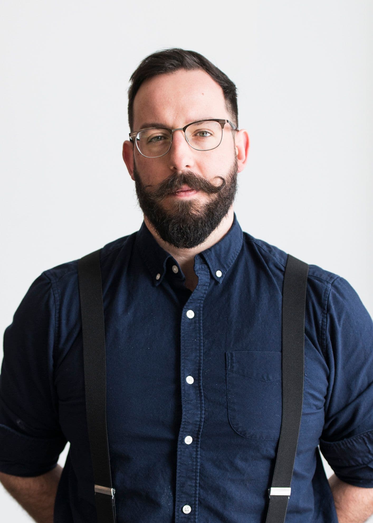 Remington, a white man, wears wire glasses, a blue button up and suspenders. He stand in front of a white background with short brown hair and a handlebar moustache.