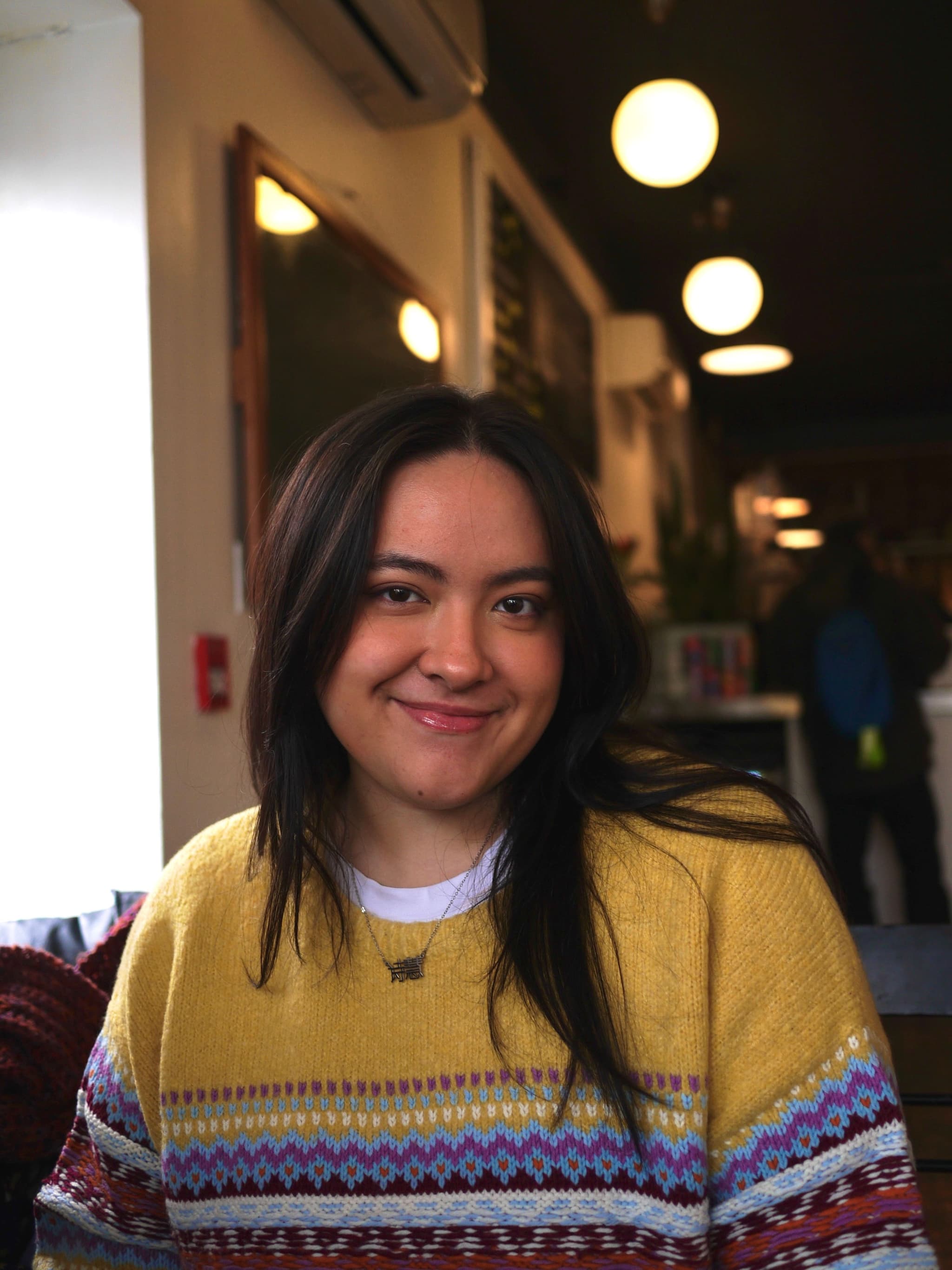 Annasofie, a mixed race woman with long brown hair, smiles in a photo. She's wearing a yellow sweater with different patterns and a white shirt underneath, with a blurred background.