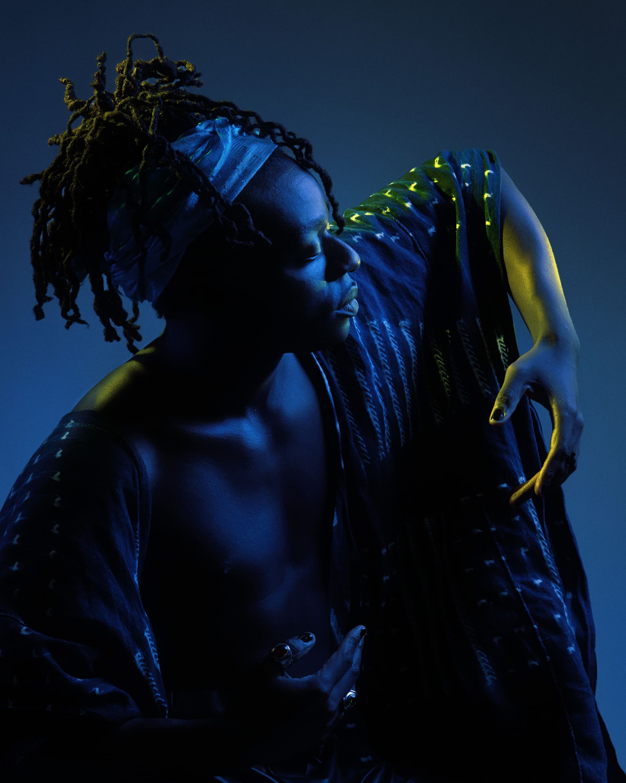 Kwasi Obeng-Adjei, a Black African-Canadian person with long dreadlocks in an artistic pose in a profiled position. He is wearing a patterned robe and a head wrap, with their hair tied up. His eyes are closed, and he is posed gracefully with one arm lifted and the other near their chest.